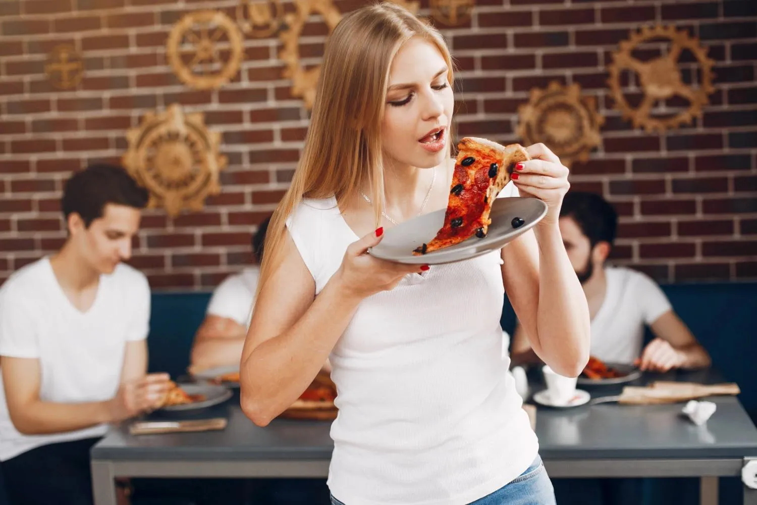 dívka se cpe pizzou, za ní sedí parta lidí u stolu v restauraci