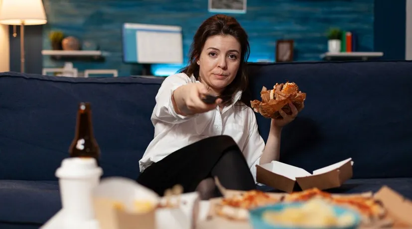 žena v jedné ruce drží ovladač, ve druhé burger a jde si pustit na gauči něco na TV