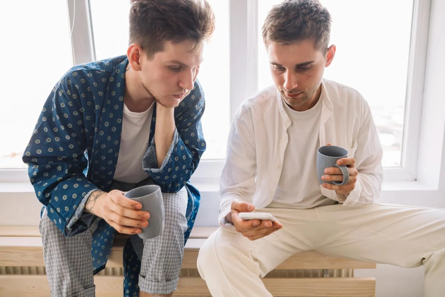 Dva mladí muži s hrnky sedí u okna a soustředěně sledují displej mobilu